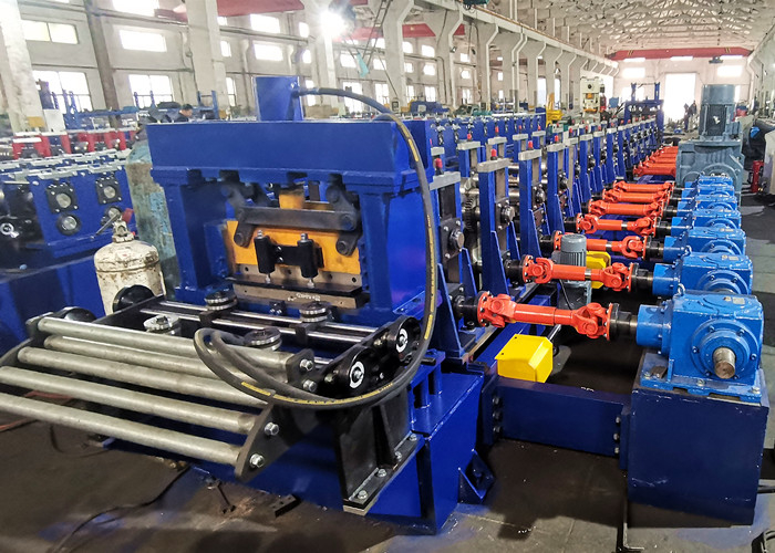 A long, blue industrial roll forming machine with numerous sets of rollers, feeding a flat sheet of metal into its sequence of shaping dies.